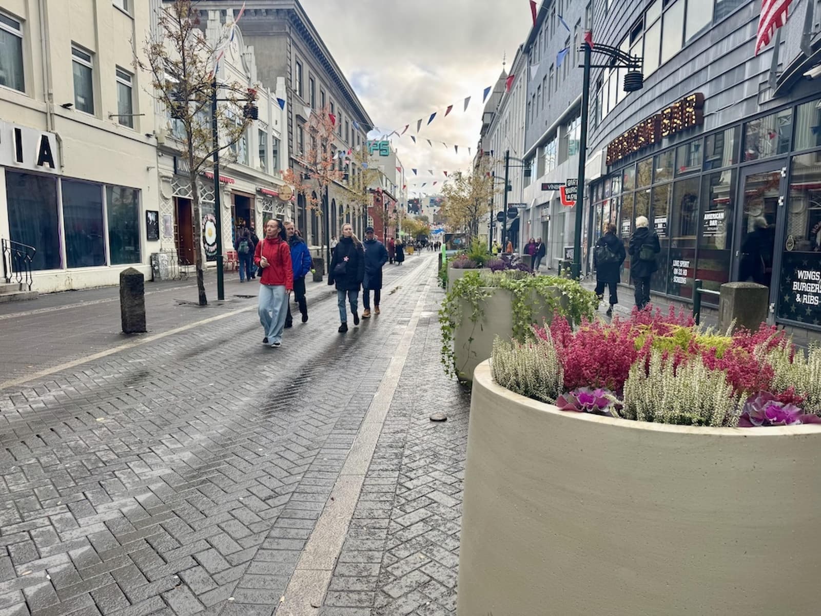 Pedestrians walking along Austurstræti in central Reykjavík beside planters and storefronts.