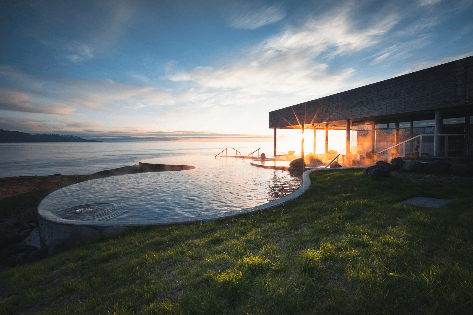 Steam rising from GeoSea's outdoor pools beside the bathhouse and Skjálfandi Bay at low sun.