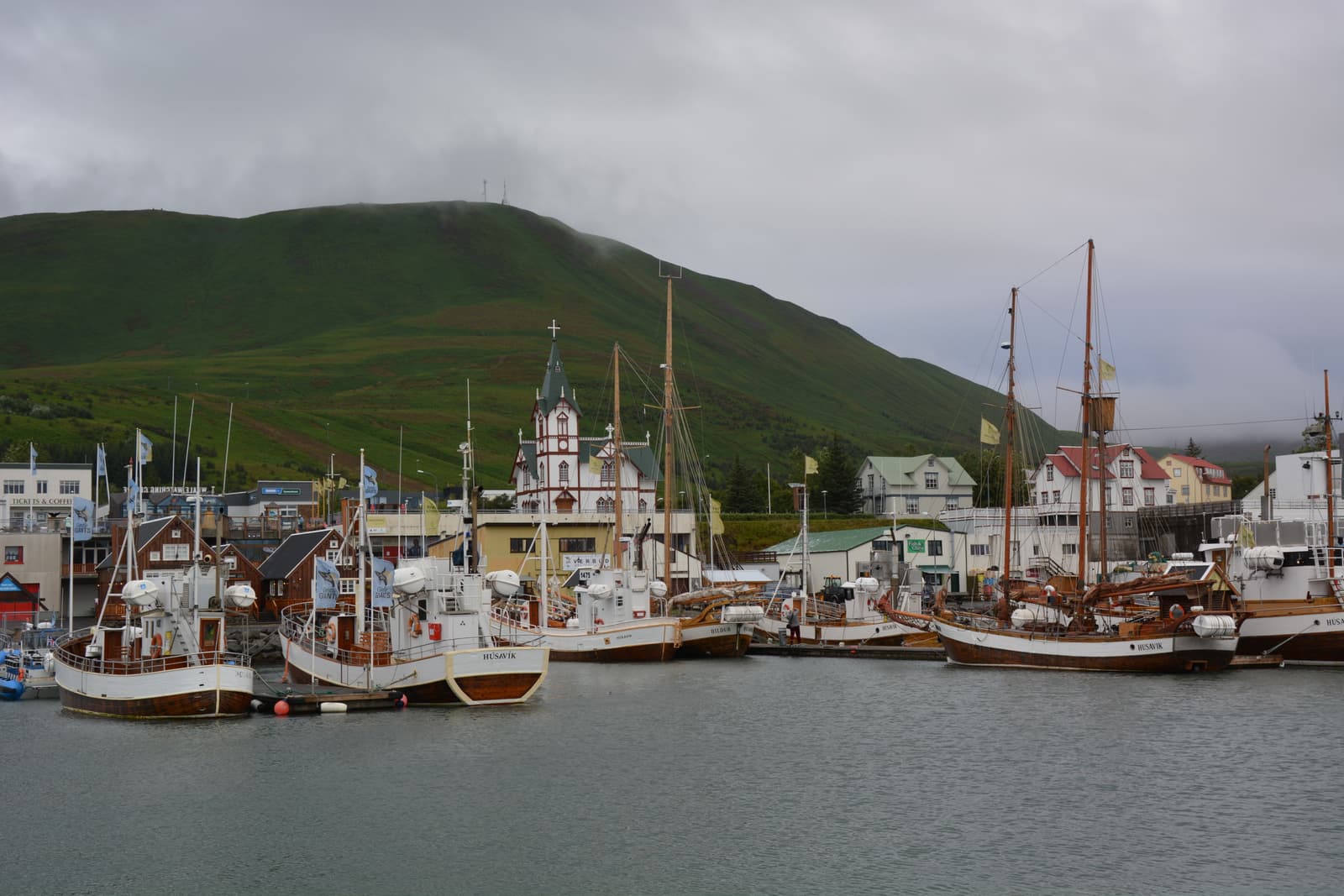 Whale-watching boats in Húsavík harbor with Húsavíkurkirkja below a misty green mountain.