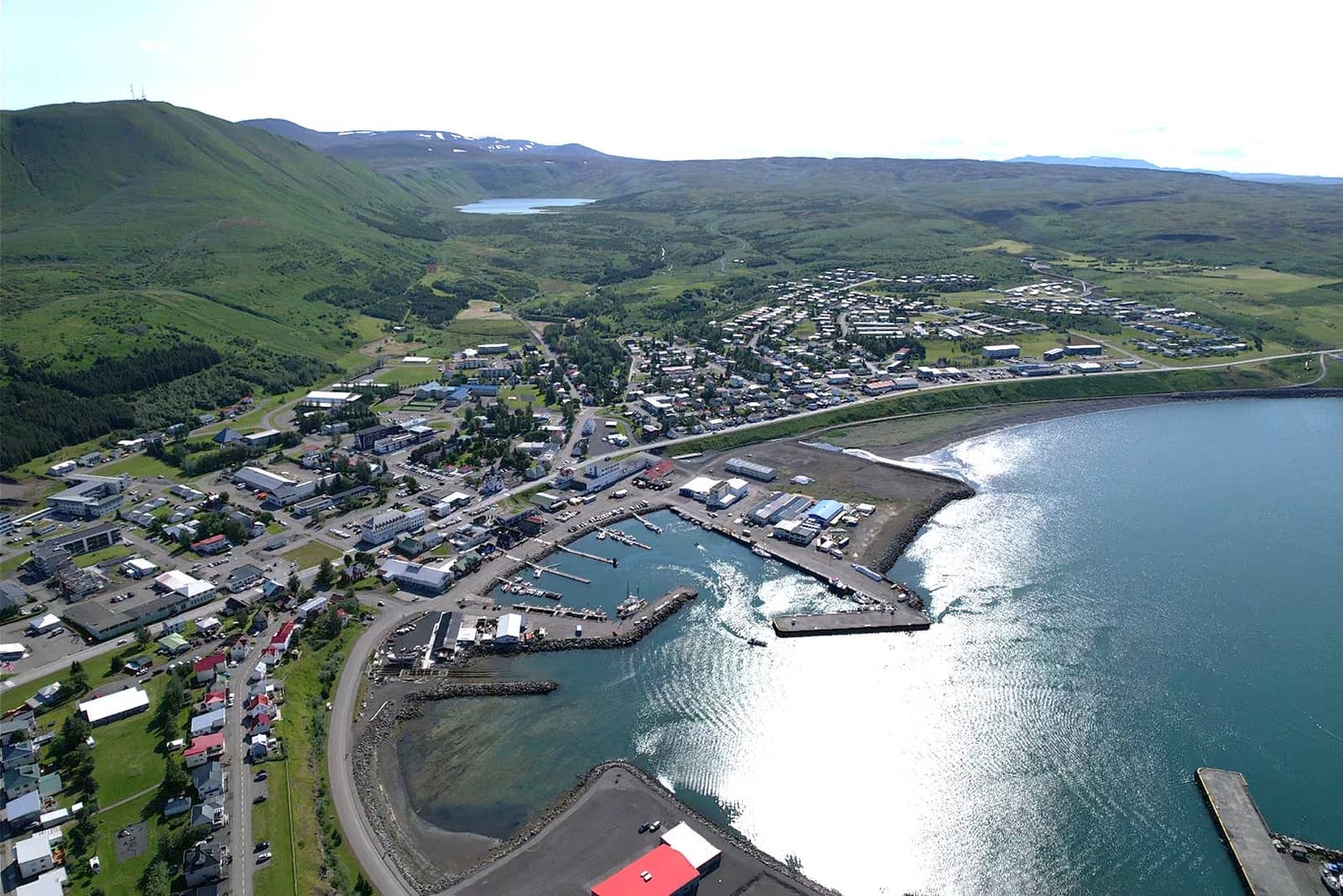 Aerial view over Húsavík harbor, town streets, and Skjálfandi Bay on a clear summer day.