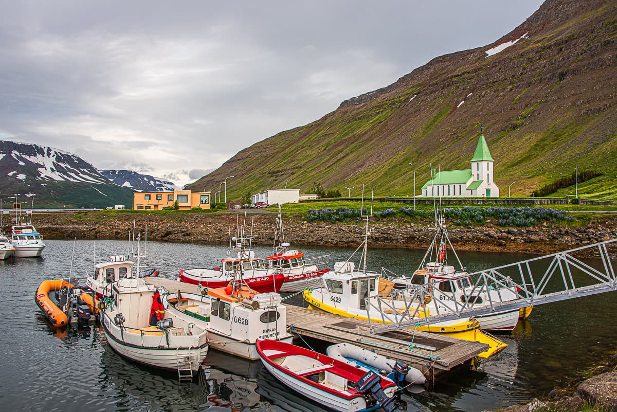 Small boats in Suðureyri harbor with the church and mountain slope beyond the quay.