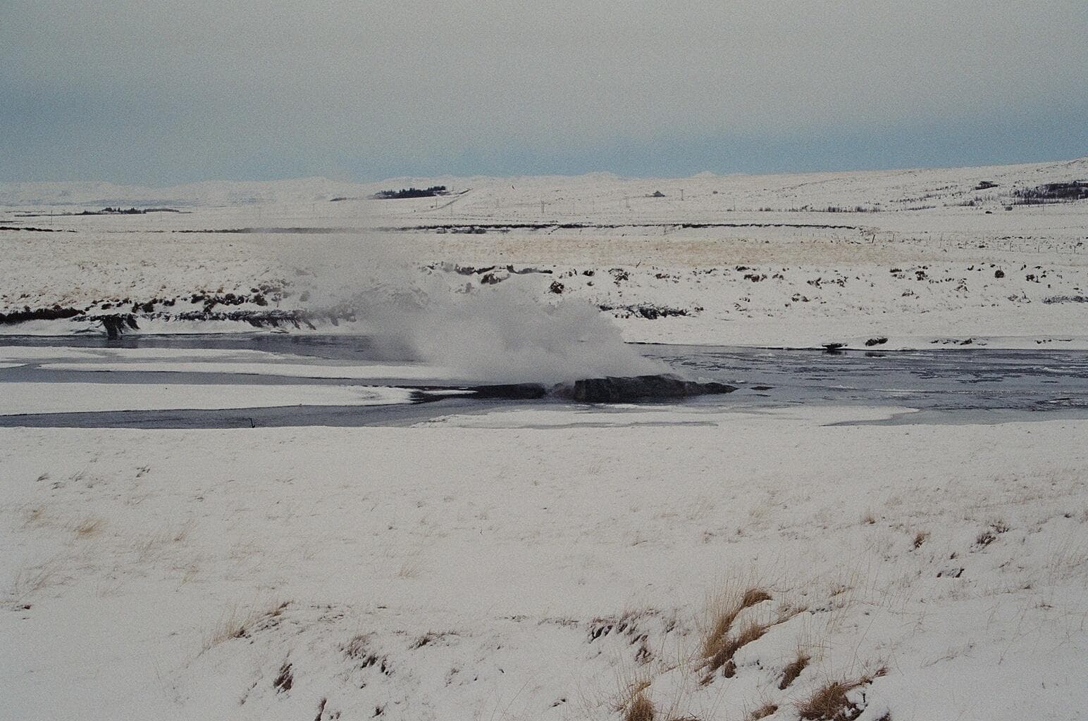 Steam rising from Árhver hot spring in the Reykjadalsá river with snowy Reykholtsdalur hills behind it.