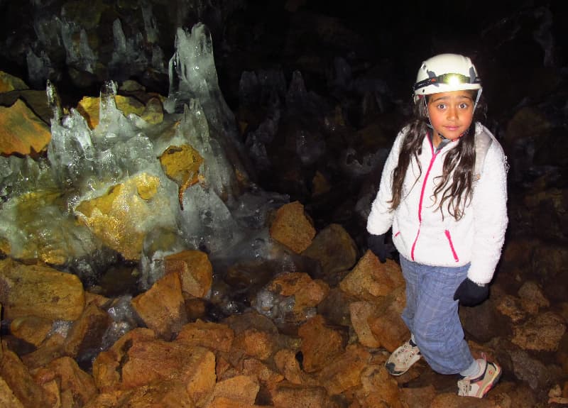 A helmeted visitor standing beside clear ice formations and loose lava rocks inside Búri Lava Cave.