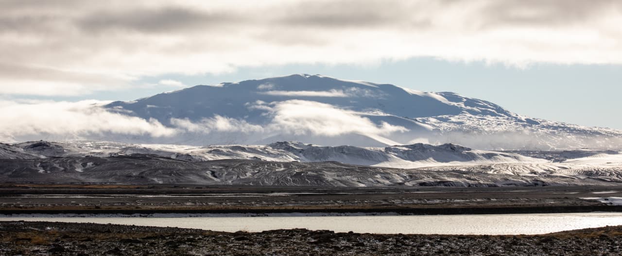 Snowy Hekla ridge seen beyond low volcanic hills and a river in South Iceland.