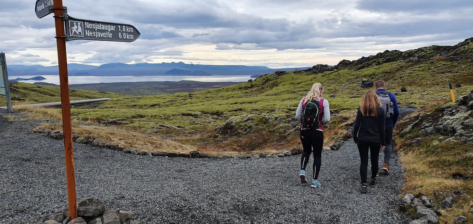 Walkers following a marked Hengill trail with a signpost, lava slopes, and Þingvallavatn in the distance.