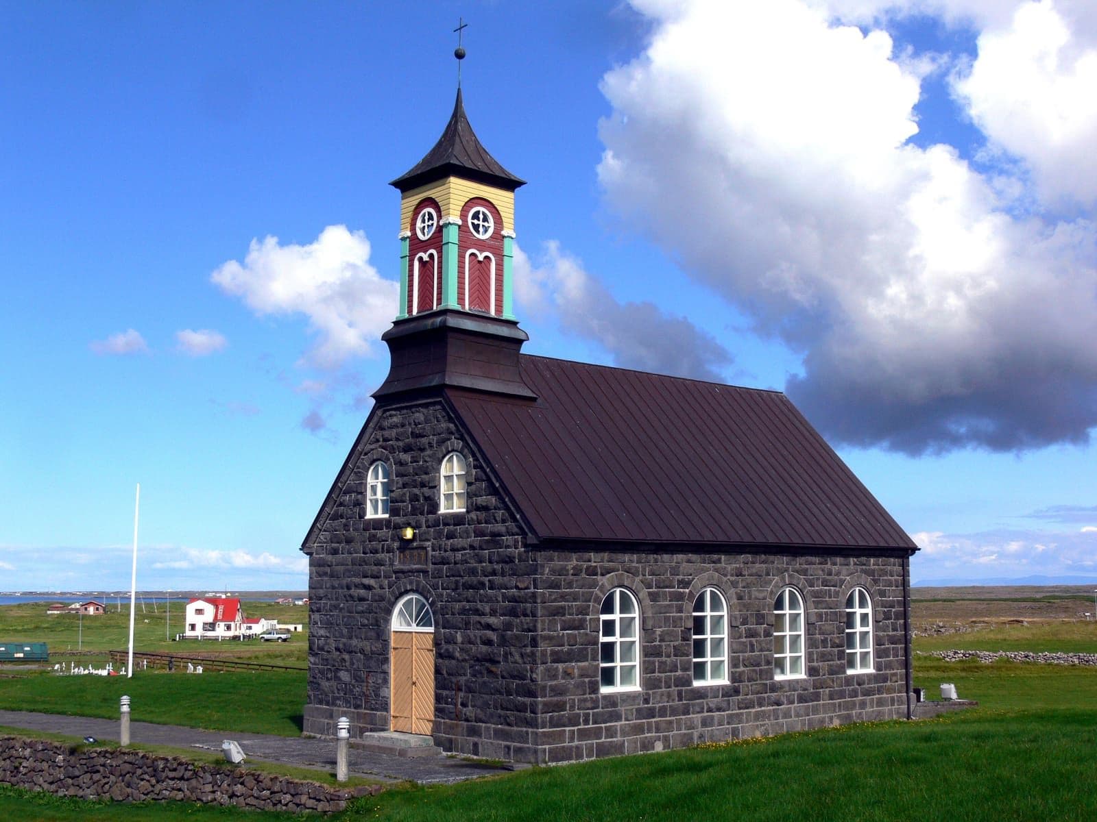 Side view of Hvalsneskirkja with its red tower, dark stone walls, and low churchyard wall.