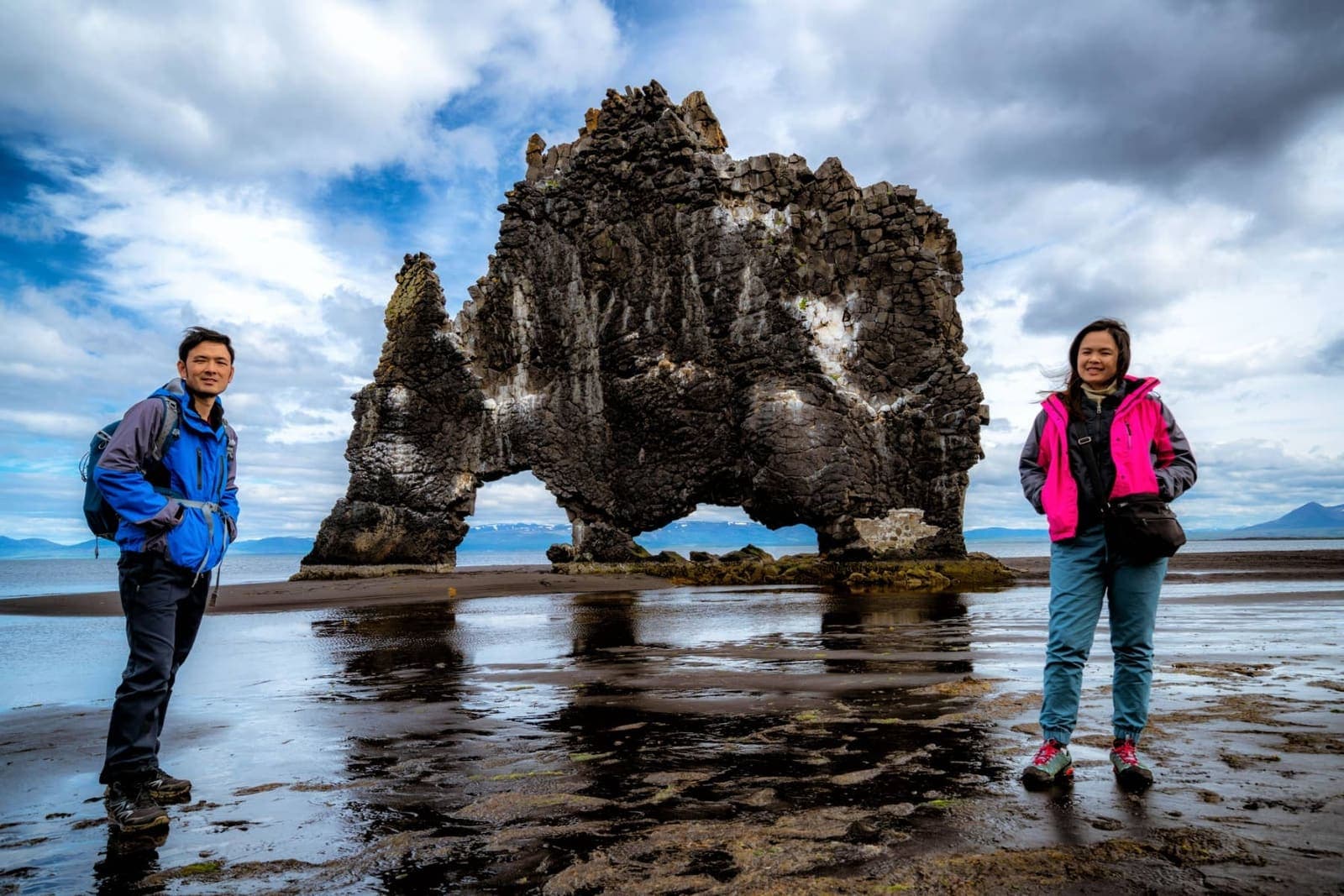 Two visitors standing on wet sand near Hvítserkur sea stack.