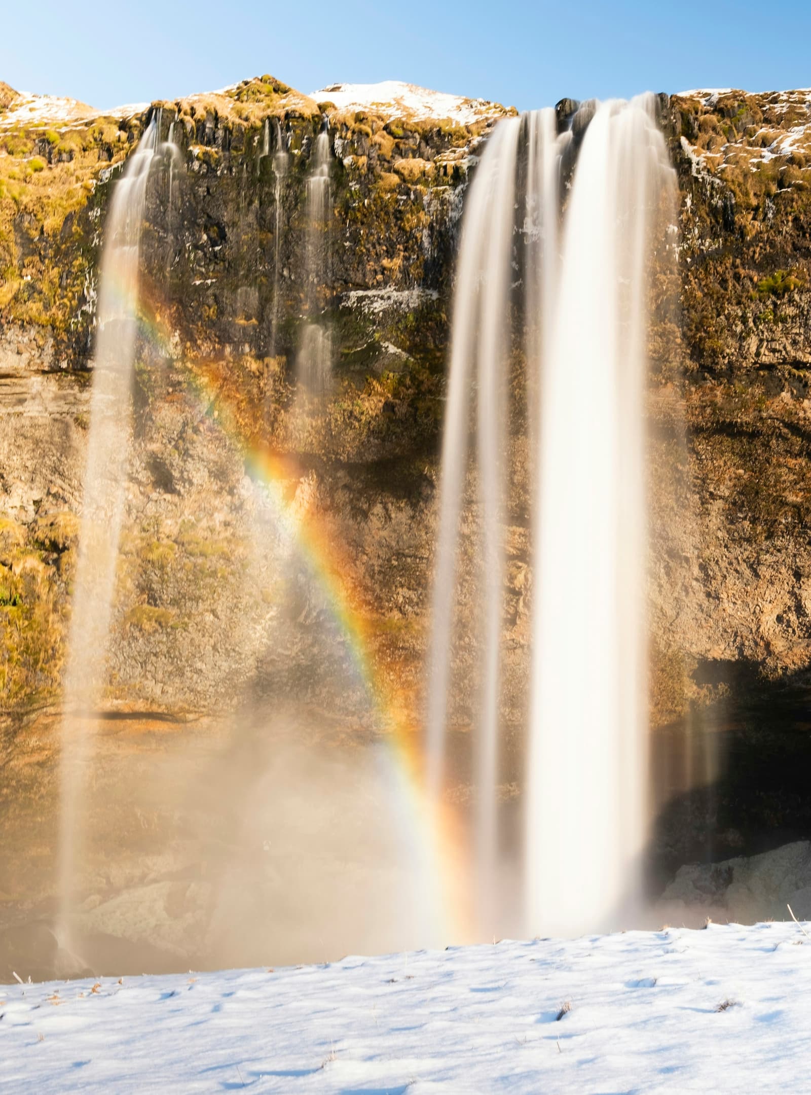 Rainbow and snow around Írárfoss waterfall in a narrow South Iceland gorge.