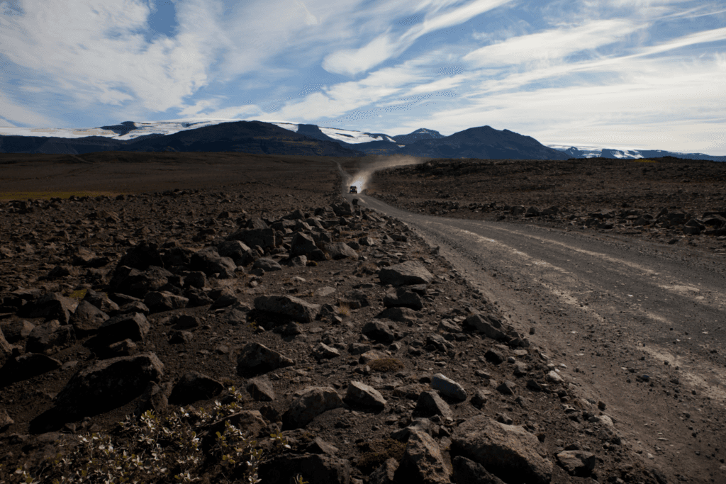 A gravel road crossing the rocky Kaldidalur valley with a vehicle dust trail and glacier views ahead.