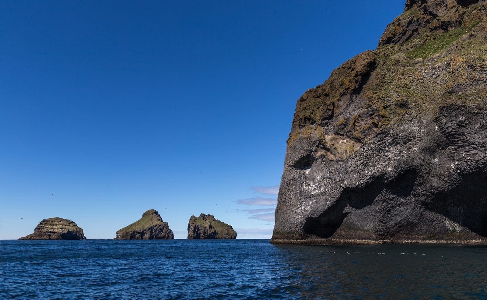 Steep Westman Islands sea cliffs and offshore rocks seen from a boat on blue water.
