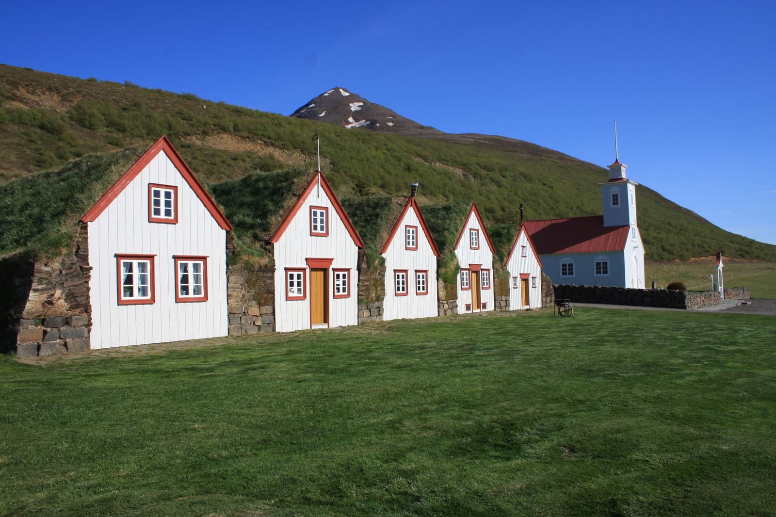 Laufás turf farm with white gables, turf roofs, church, green hillside, and mountain in Eyjafjörður.