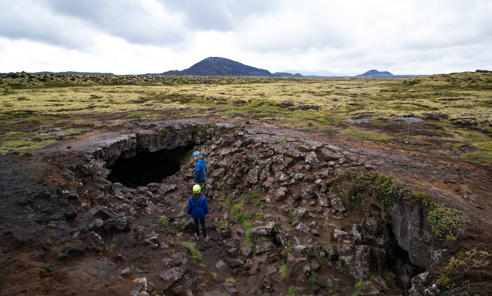 Two helmeted visitors standing by the rocky Leiðarendi cave entrance in a mossy lava field near Bláfjöll.