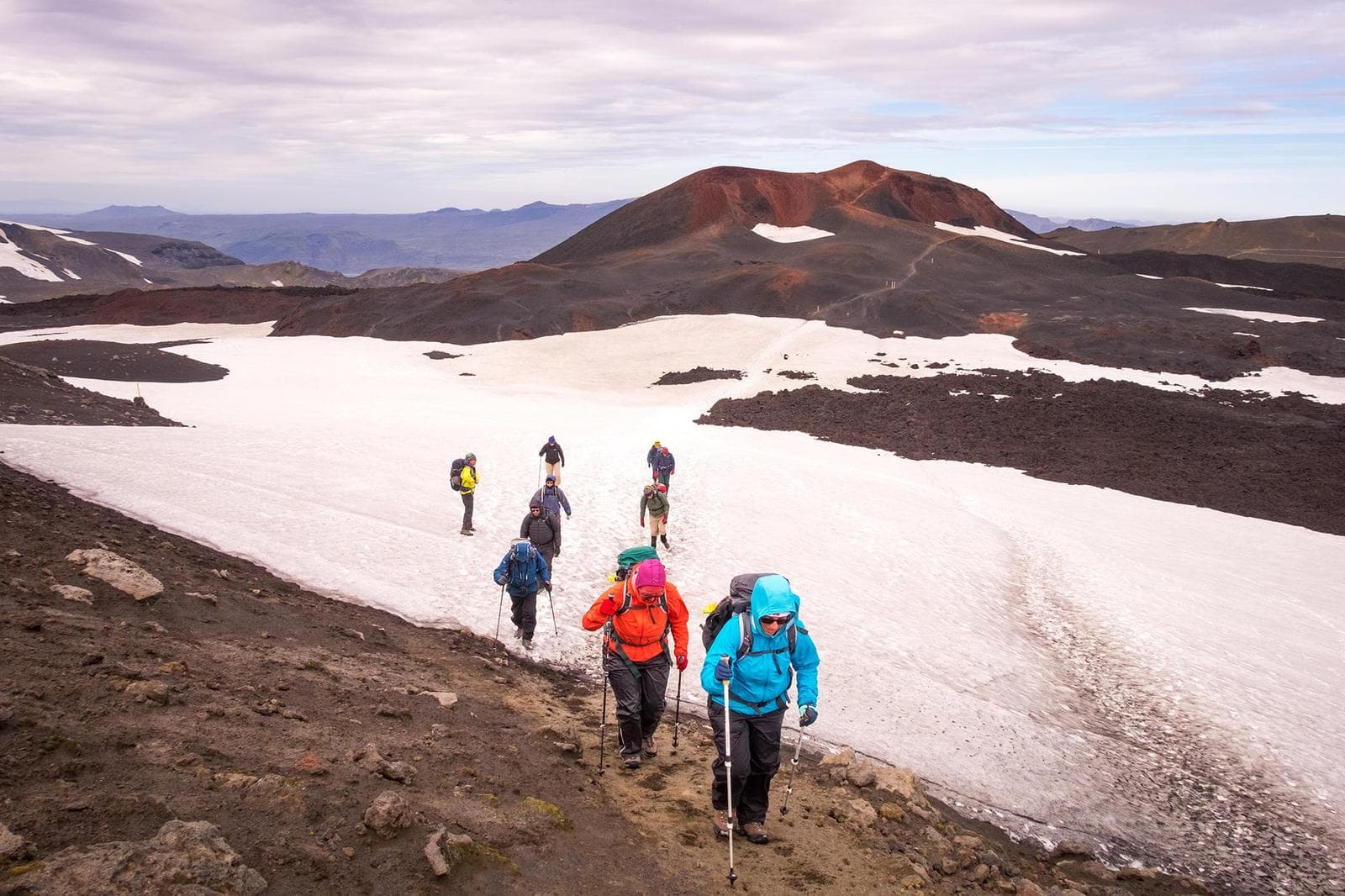Hikers crossing snow below the red Móði and Magni crater terrain on Fimmvörðuháls.