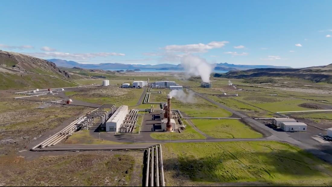 Nesjavellir power station with steam rising from pipes and buildings below green hills.