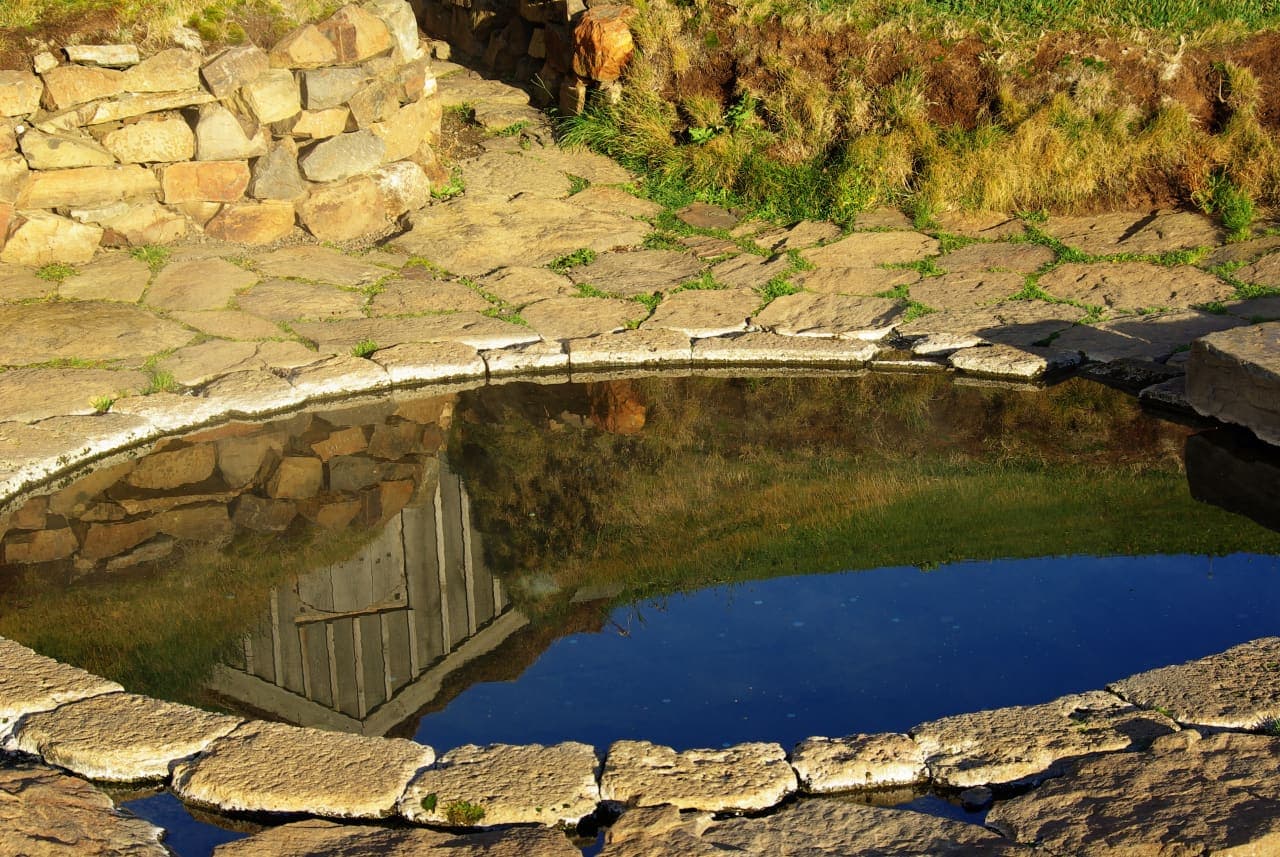 Stone edge and still water of Snorralaug reflecting the small turf-covered passage house