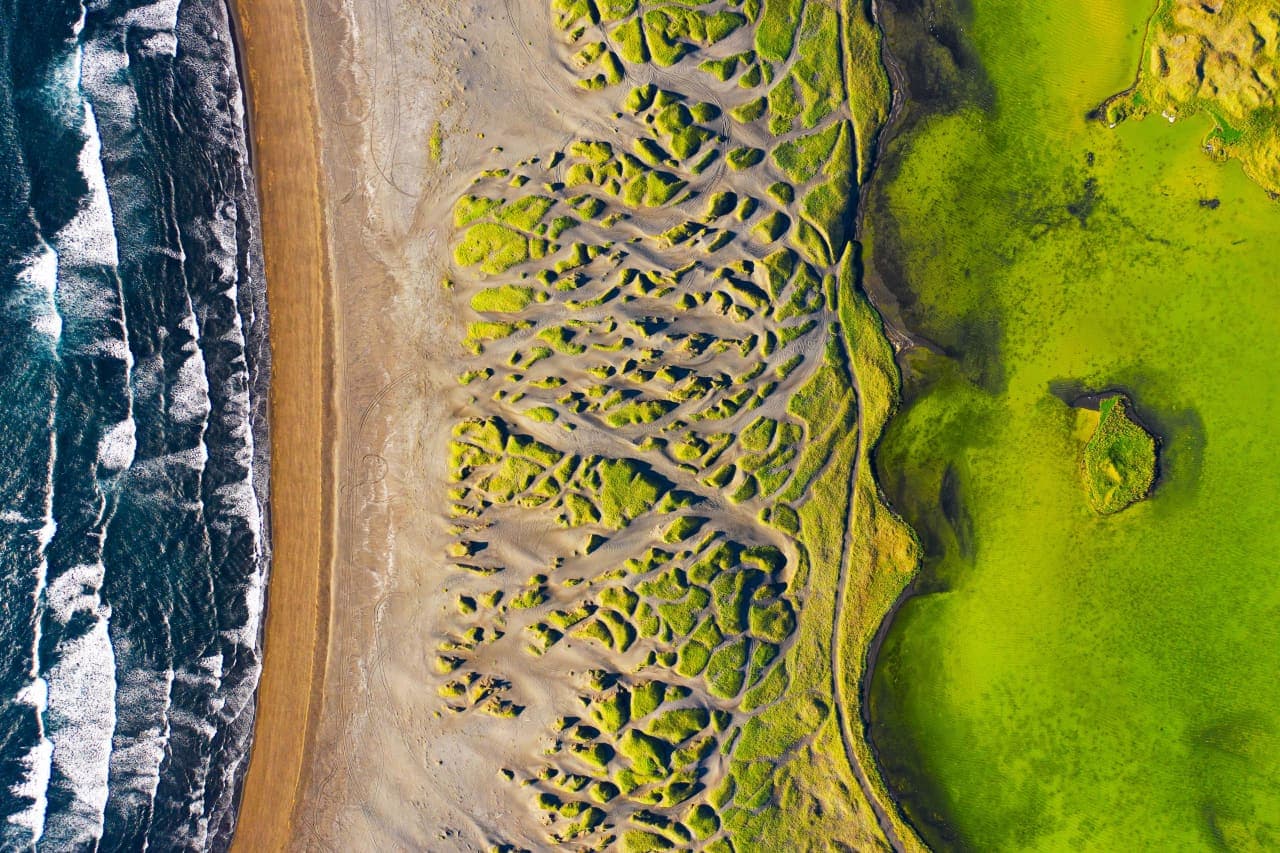 Aerial view of Sandvík with black sand beach, grassy dunes, pond water, and Atlantic surf on Reykjanes.