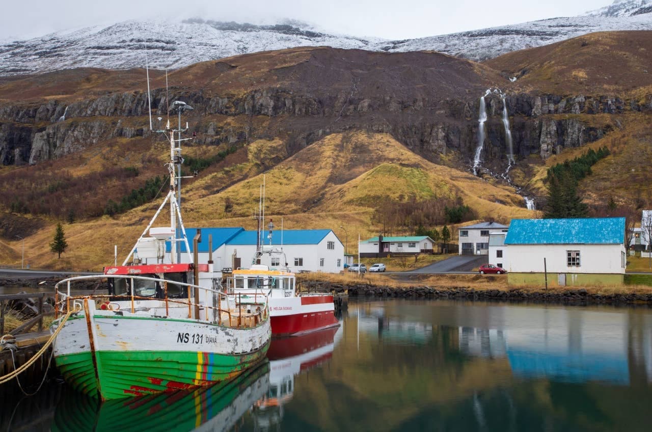 Fishing boats in Seyðisfjörður harbor with waterfalls and mountains behind the Eastfjords town.