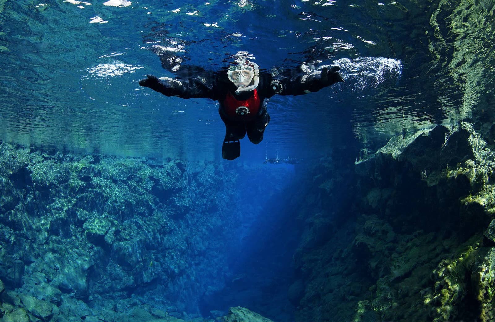 Snorkeler floating between dark lava walls in the clear blue water of Silfra fissure.