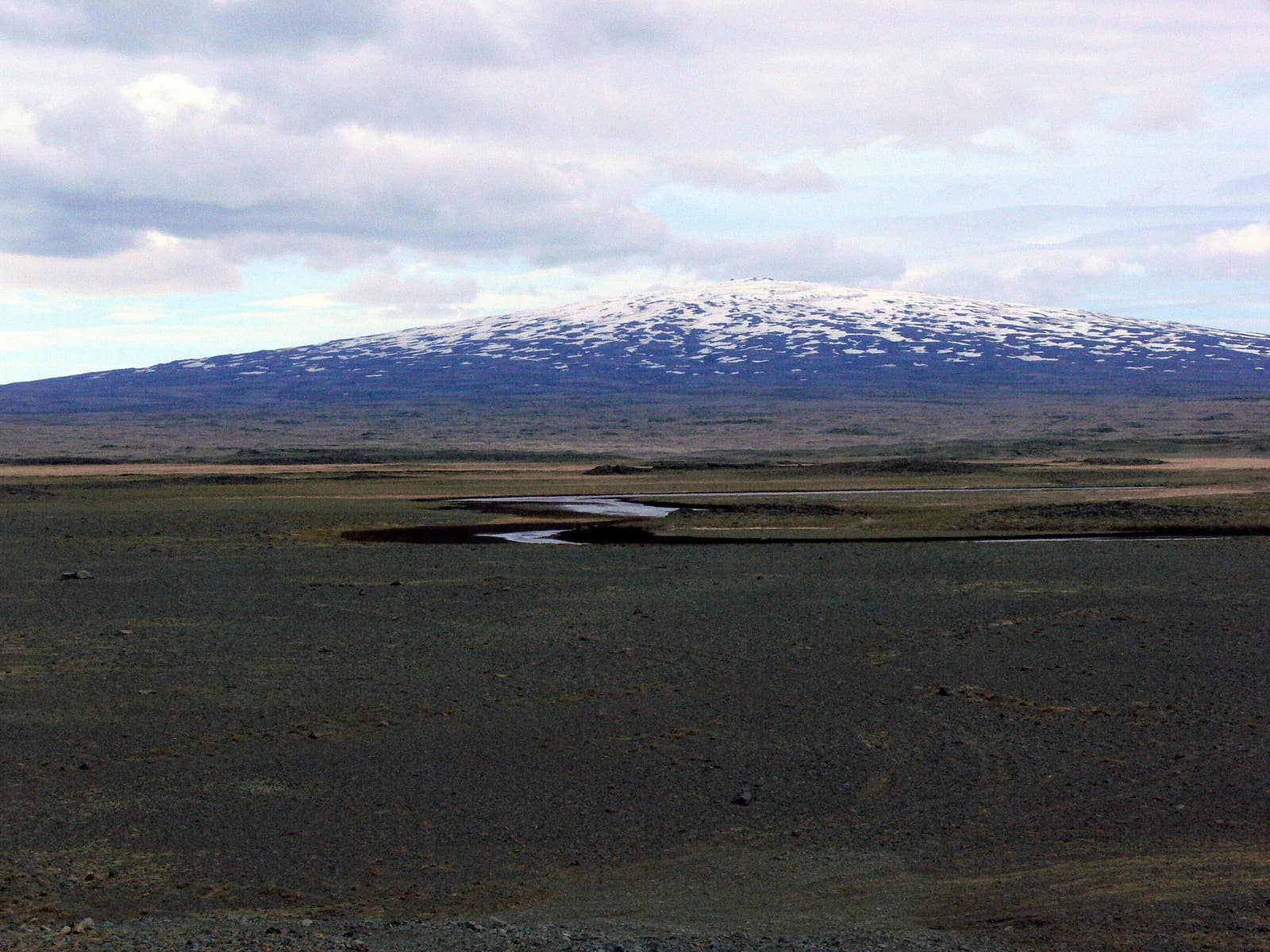 Skjaldbreiður with a patchy snow cap rising beyond dark lava ground and shallow water.