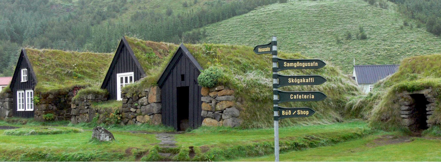 Turf-roofed buildings and visitor signs at Skógar Museum below green South Coast slopes.