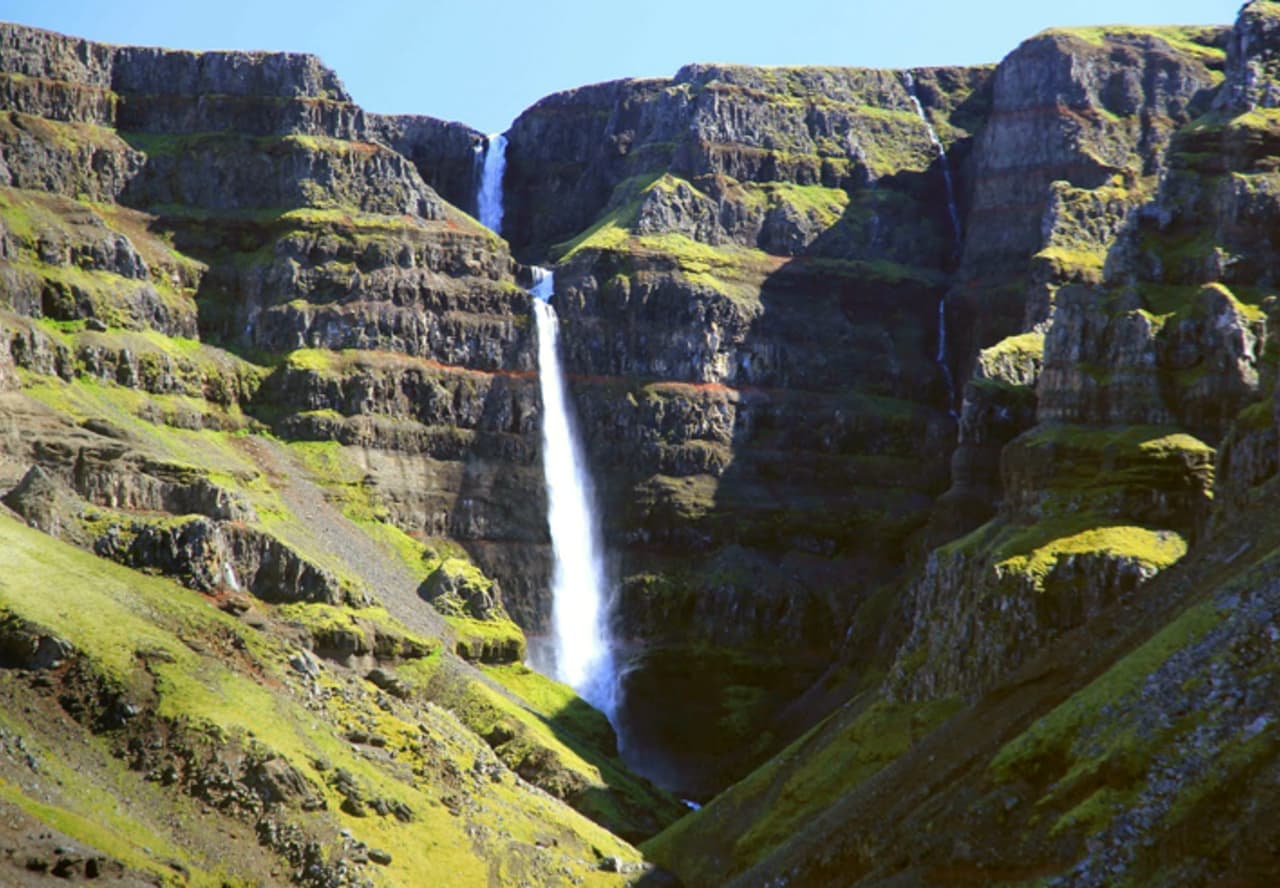 Strútsfoss dropping in two main tiers through a deep East Iceland cliff valley with red and dark rock bands.