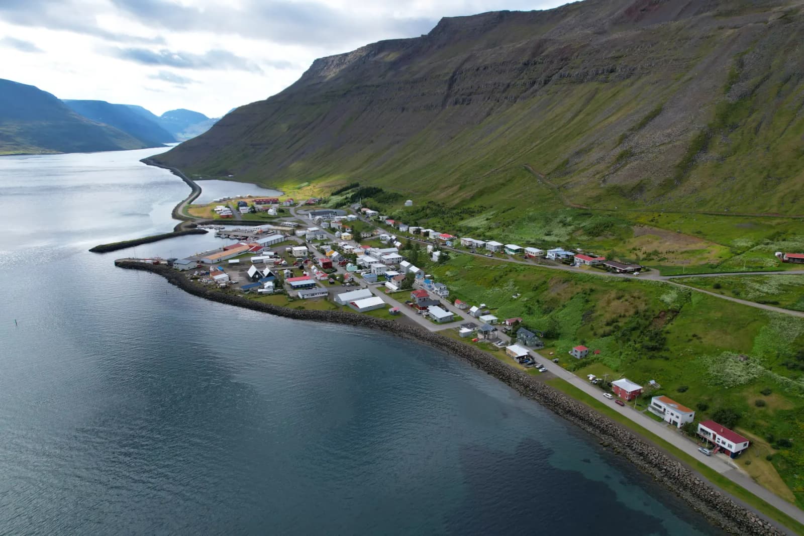 Aerial view of Suðureyri at the mouth of Súgandafjörður with clustered houses, harbor edge, and steep mountains behind.