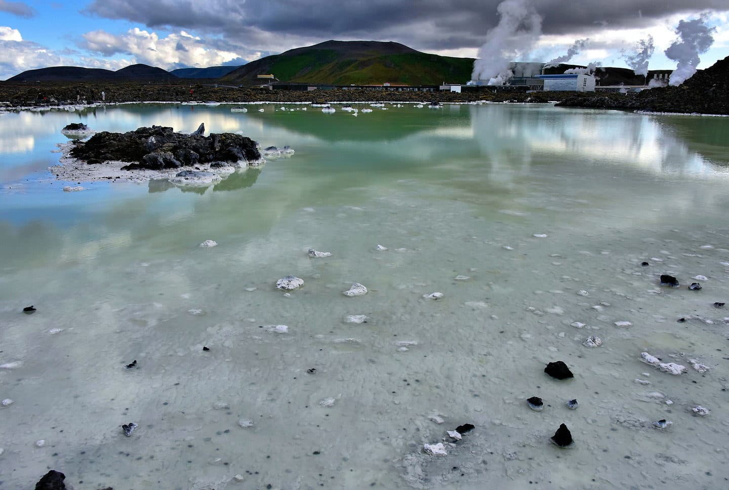 Svartsengi Power Station steaming beyond pale geothermal water and black lava on the Reykjanes Peninsula.