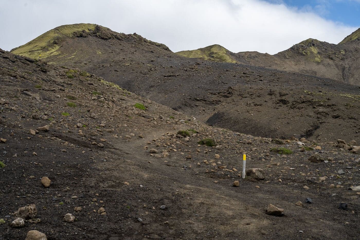 Marked Sveinstindur trail crossing loose dark volcanic ground toward low ridges.