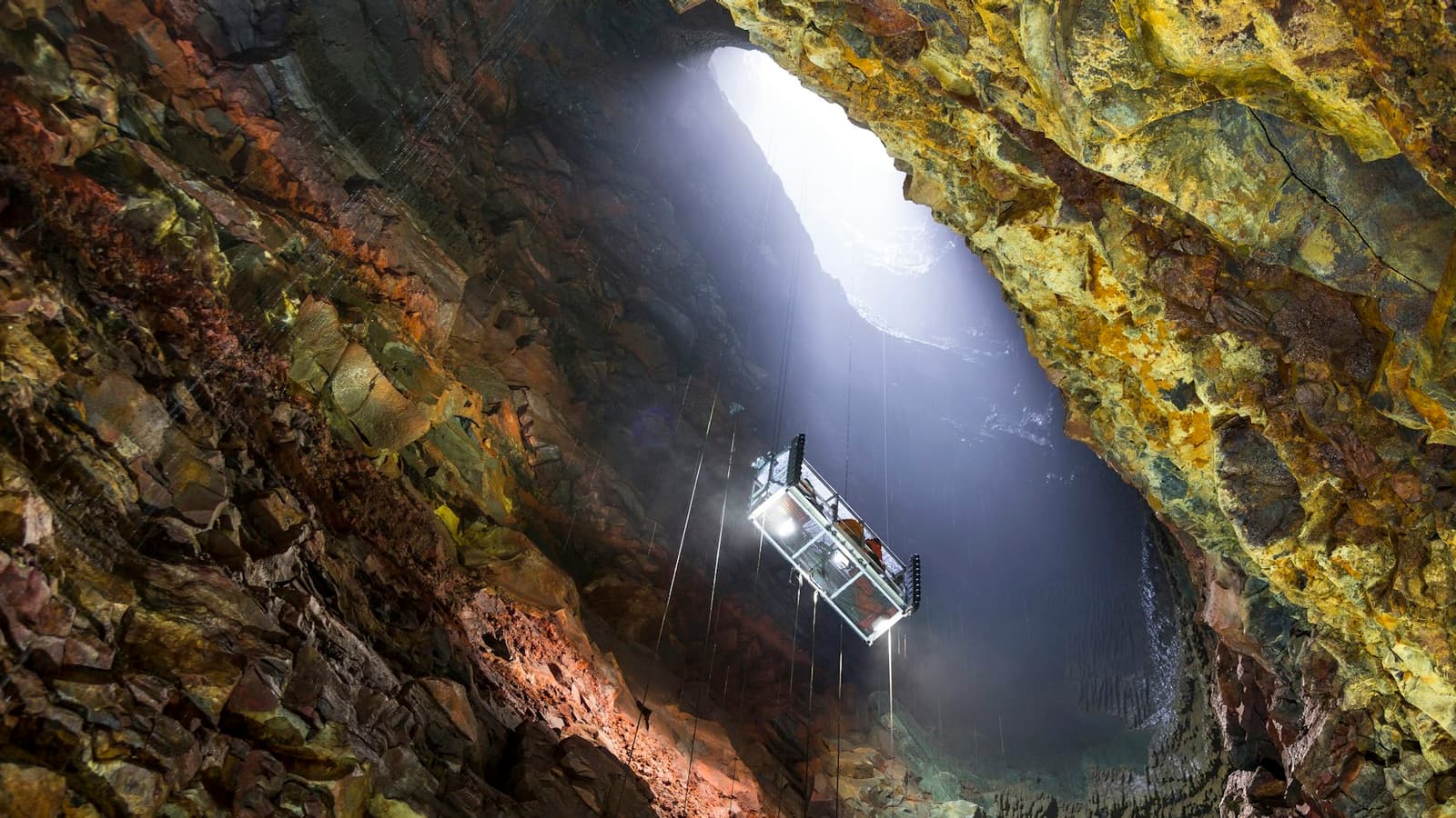Cable lift descending through the opening of Þríhnúkagígur into a broad mineral-colored magma chamber.