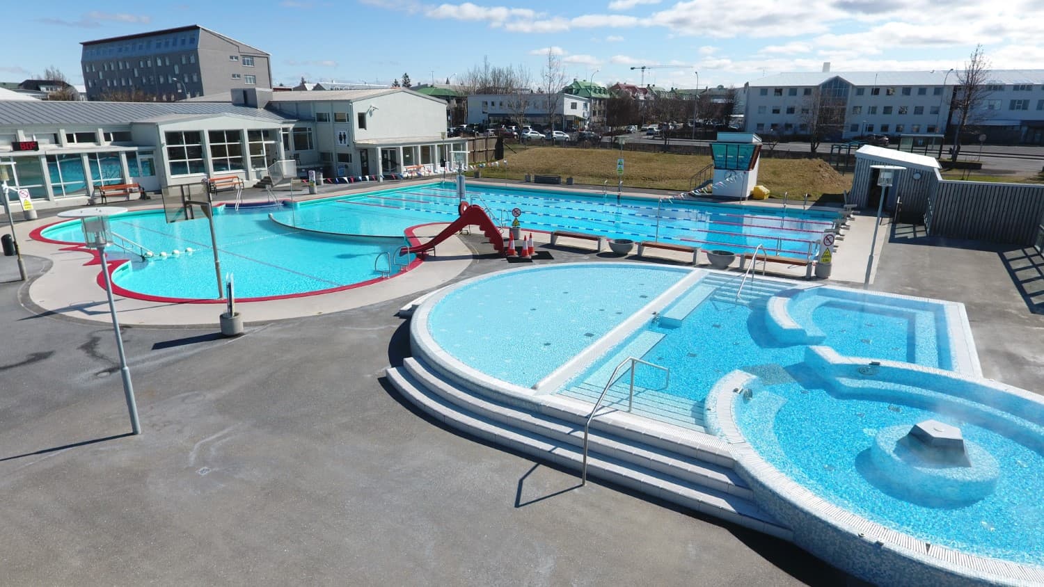 Vesturbæjarlaug outdoor pool complex with lap lanes, rounded hot tubs, children’s area, and neighborhood buildings behind the water.