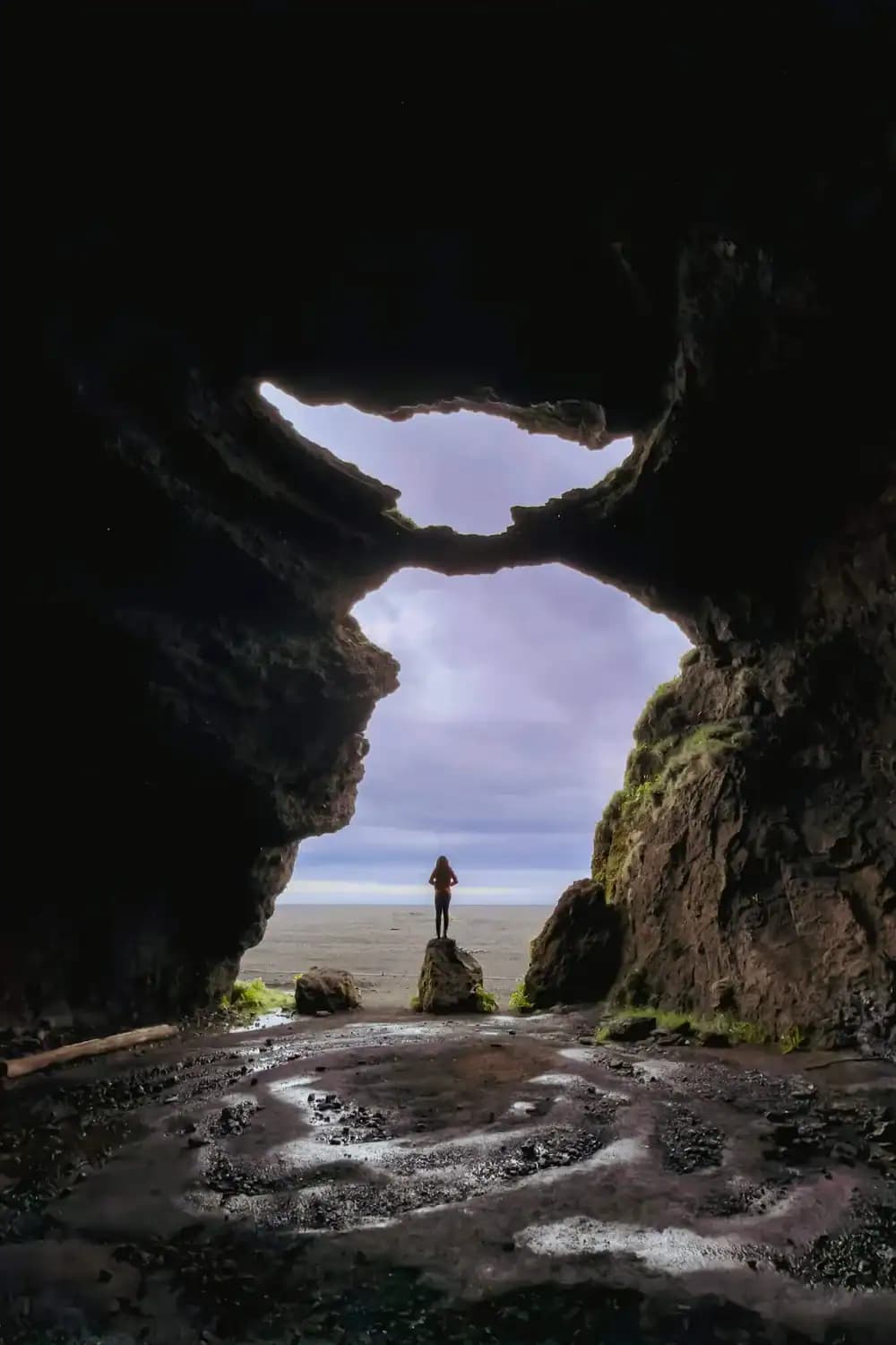 Person standing inside Gígjagjá under the Yoda-shaped cave opening at Hjörleifshöfði.