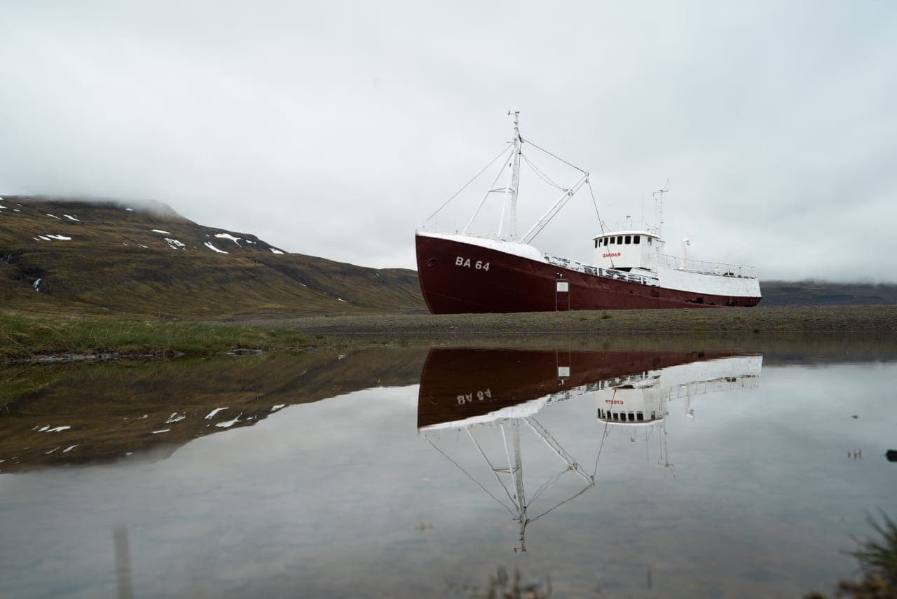 Garðar BA 64 resting on the shoreline near Patreksfjörður with its reflection in still water.