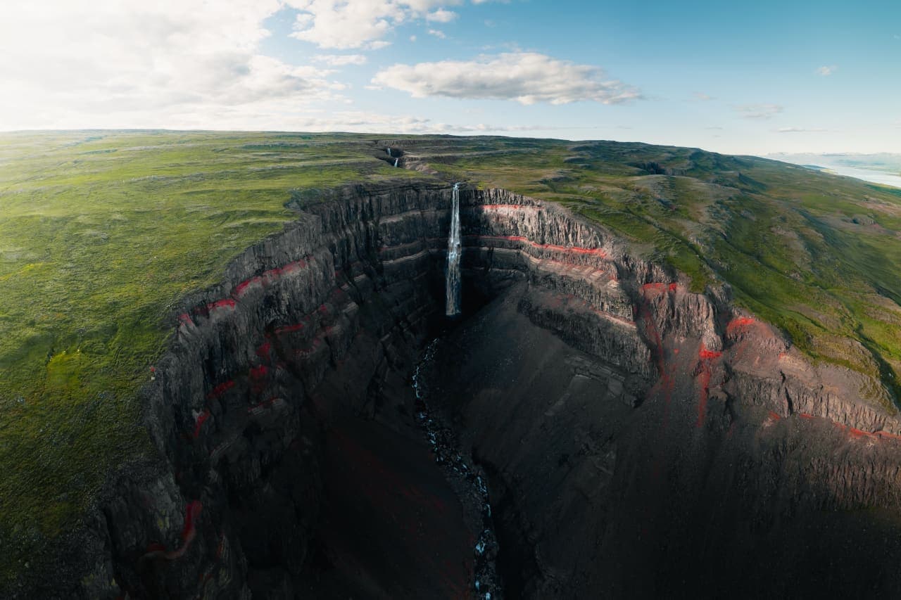 Aerial view of Hengifoss dropping through a dark basalt gorge with red sediment layers in East Iceland.