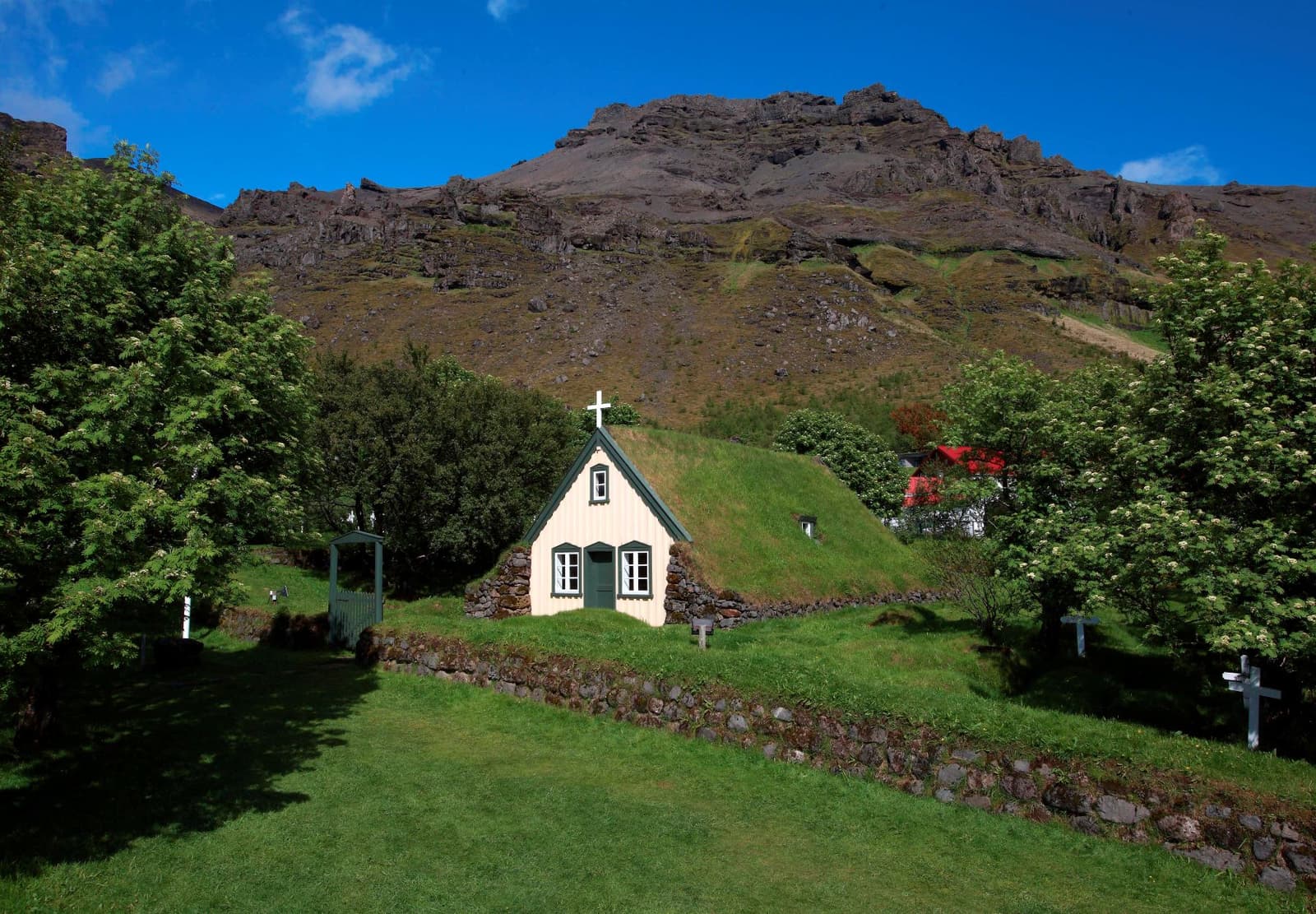 Hofskirkja Turf Church sitting inside its churchyard in Öræfi with a steep mountain wall behind it.