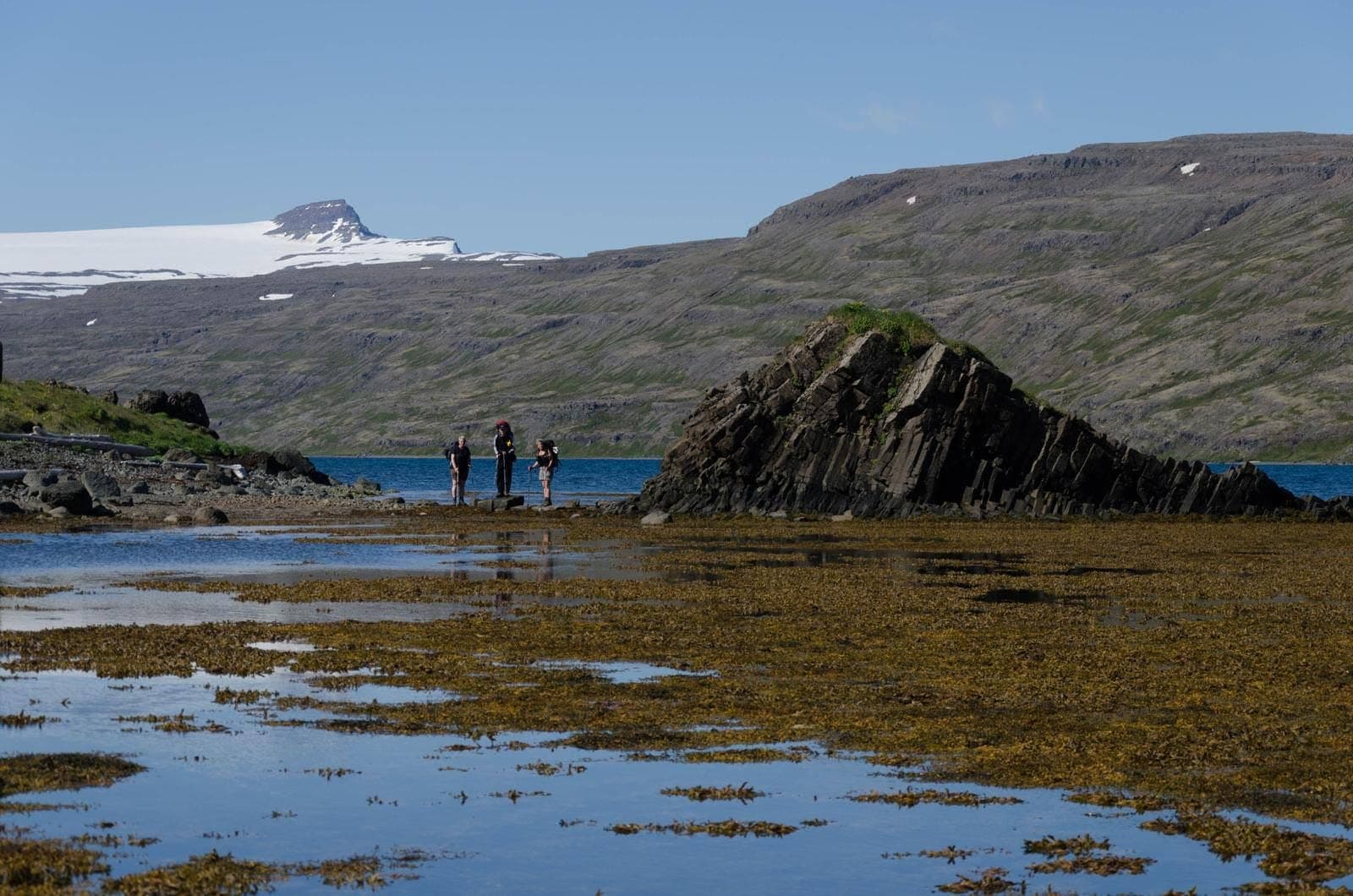 Hikers beside a rocky shore with Hrolleifsborg rising beyond water and snow patches in the Westfjords.