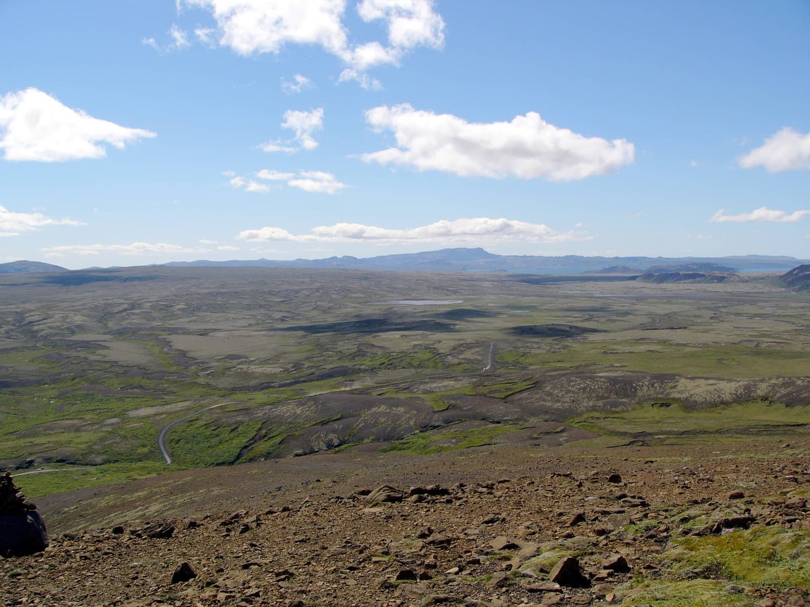 Open Lyngdalsheiði heath with dark lava ridges, a road line, low mountains, and wide blue sky.