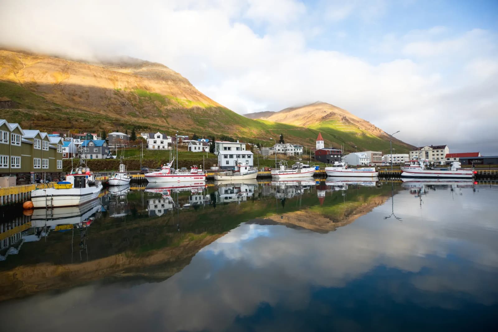 Siglufjörður harbor with fishing boats, colorful town buildings, and steep Tröllaskagi mountains above the water.