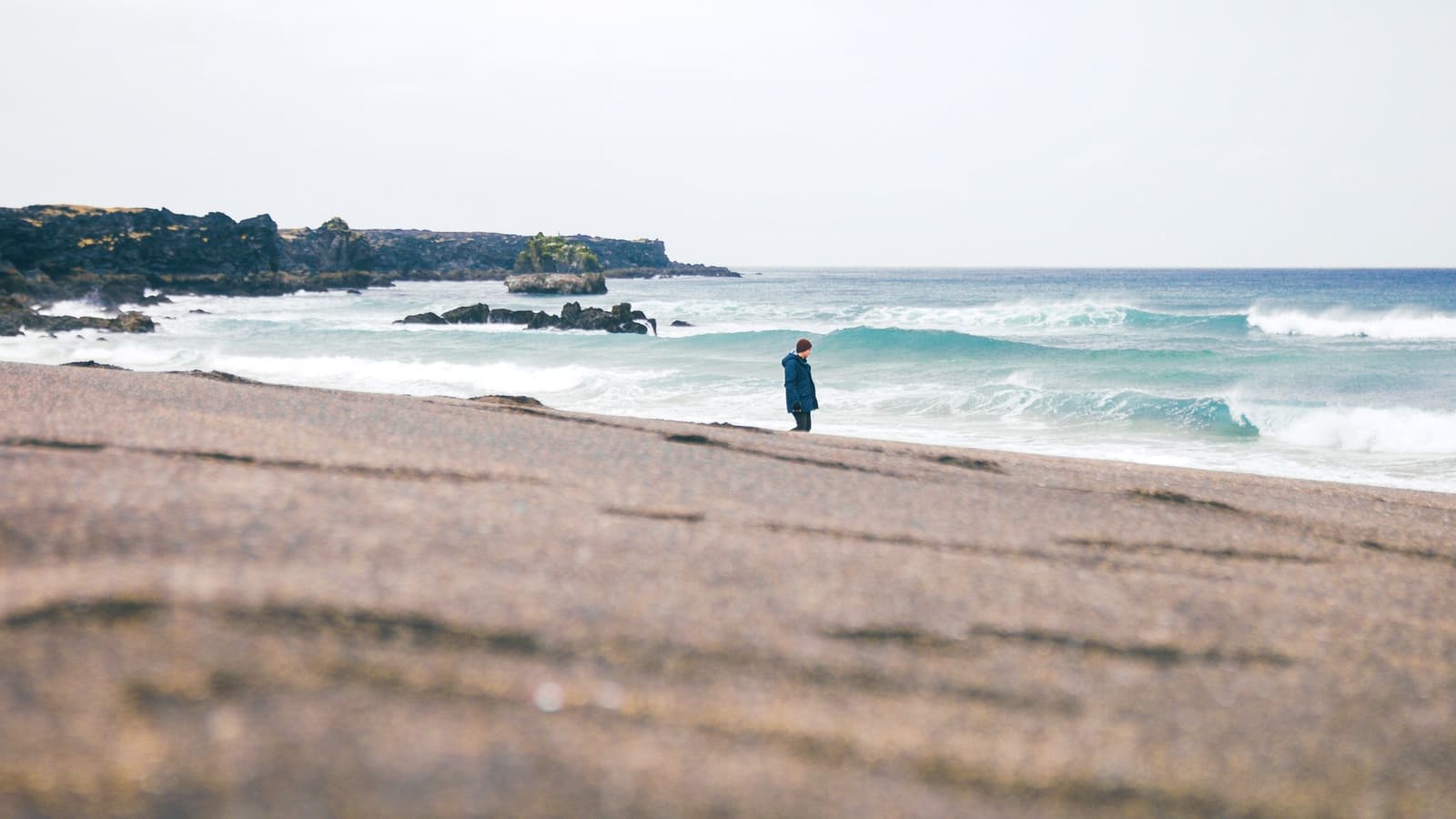 Visitor standing on Skarðsvík Beach with yellow sand, turquoise surf, and dark lava cliffs.
