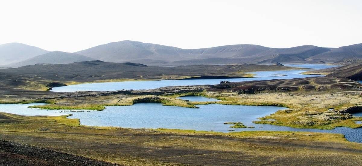 Wide view across blue Veiðivötn crater lakes, black volcanic ground, mossy islands, and low highland mountains.
