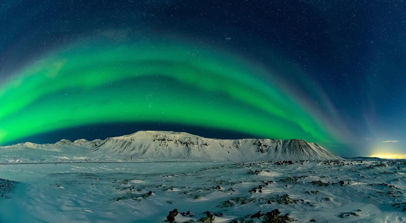 Green northern lights spreading above snowy Icelandic mountains under a starry winter sky.