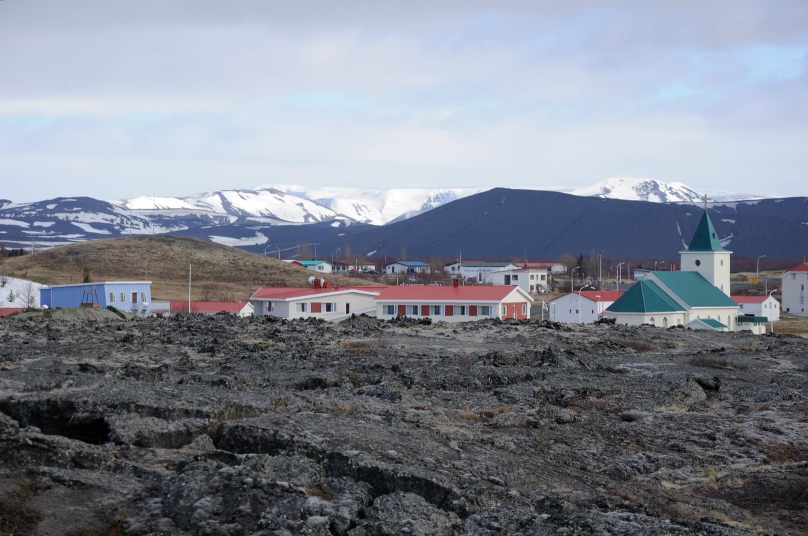 Reykjahlíð village and church seen across lava with dark volcanic slopes and snowy mountains behind.