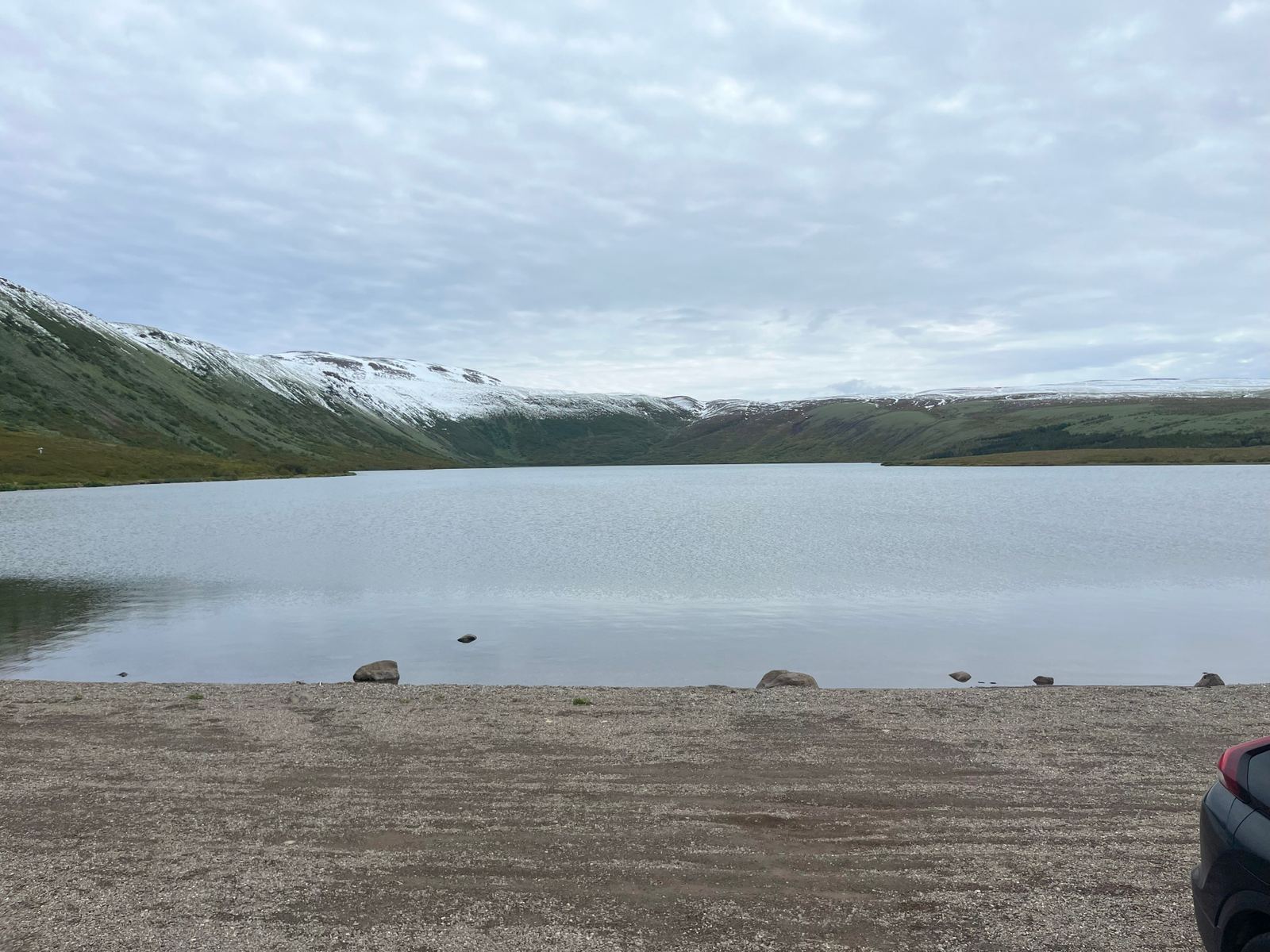 Calm Botnsvatn water and low mountain slopes above Húsavík under cloudy sky.