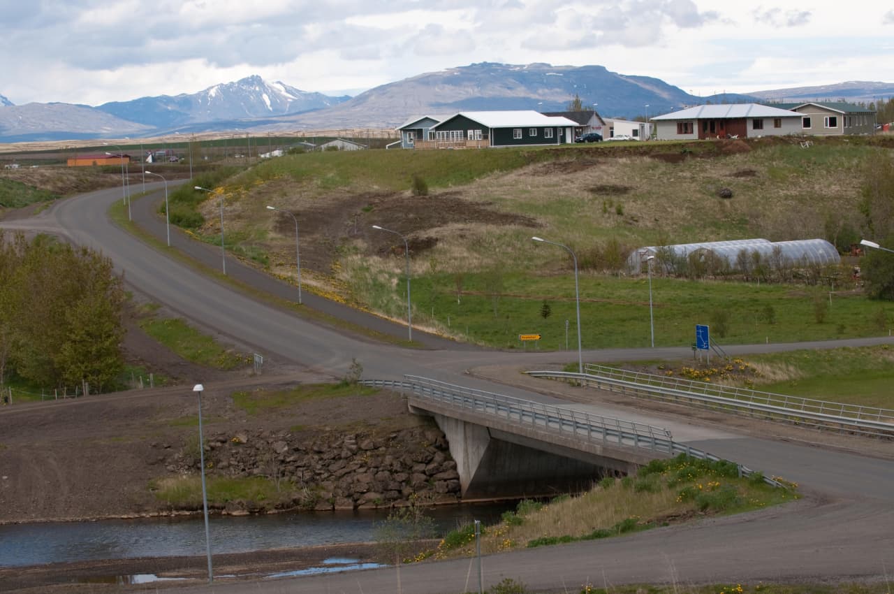 Bridge, river, and low buildings at the edge of Flúðir with mountain ridges beyond the village.