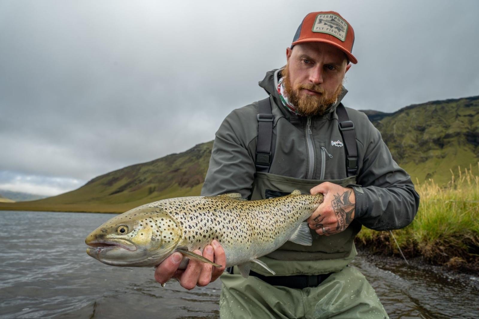 Angler holding a trout beside Lake Heiðarvatn with green slopes behind.