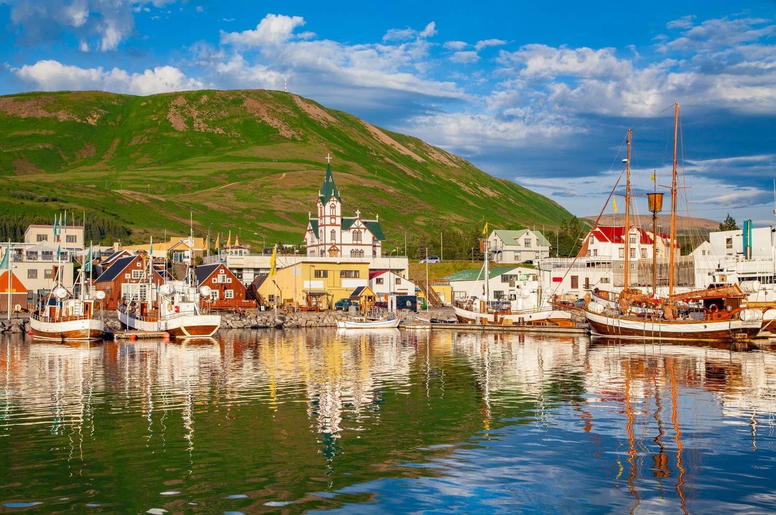 Húsavík harbor with wooden boats, Húsavíkurkirkja, and green hills above the town.