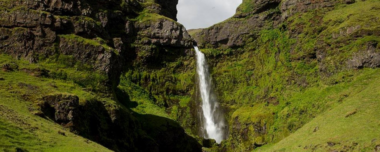 Wide view of Írárfoss falling into a green hollow below South Iceland cliffs.