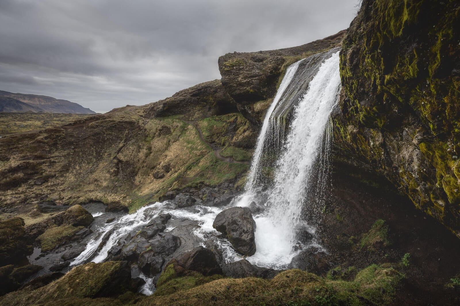 Side view of Selvallafoss falling from a mossy cliff into dark rocks below.