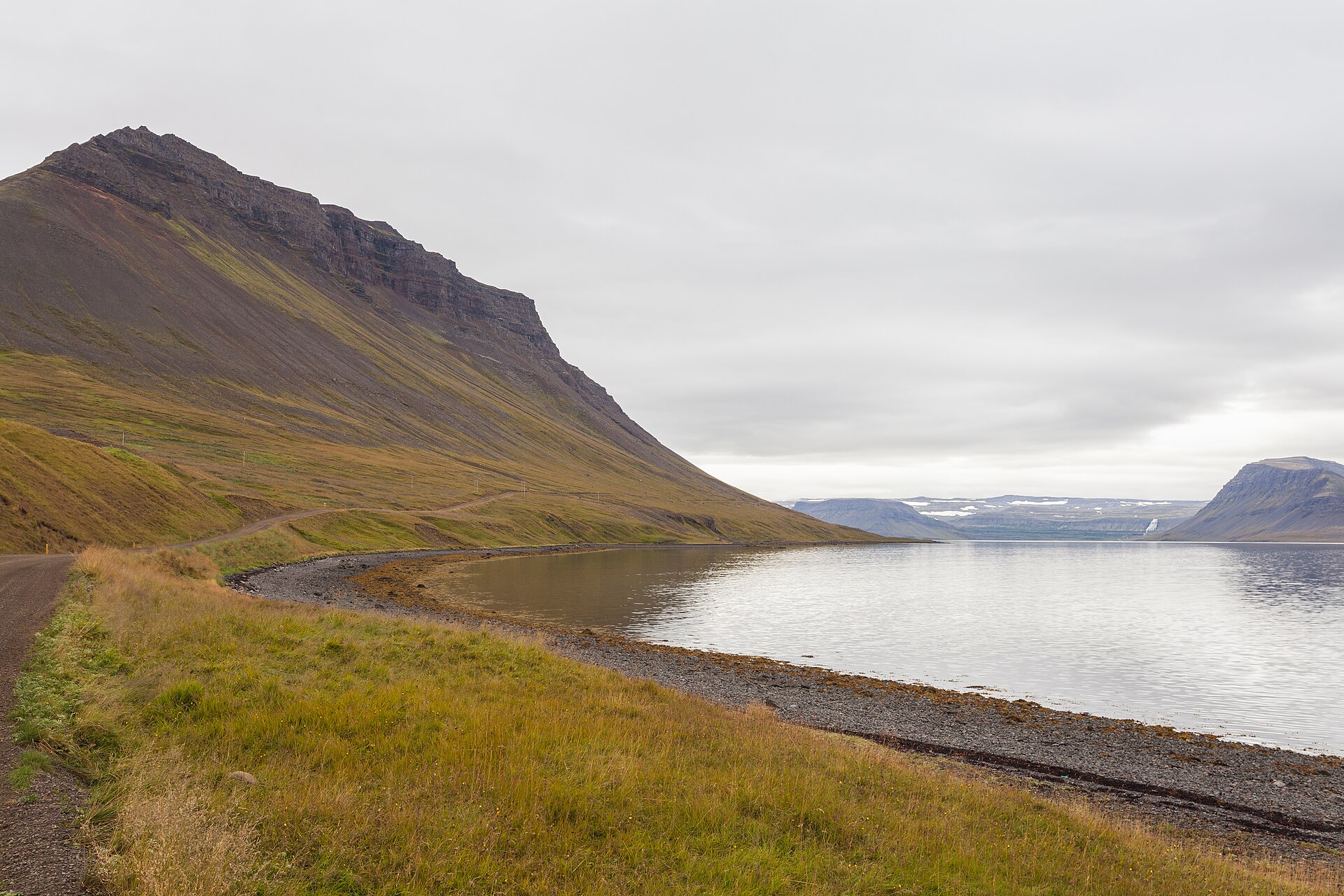 Arnarfjörður shoreline road below a steep mountain with calm fjord water and distant ridges.