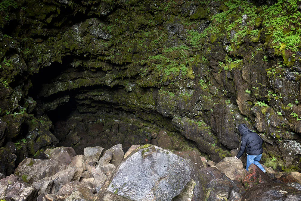 A visitor stepping over large rocks near the dark Arnarker cave entrance.