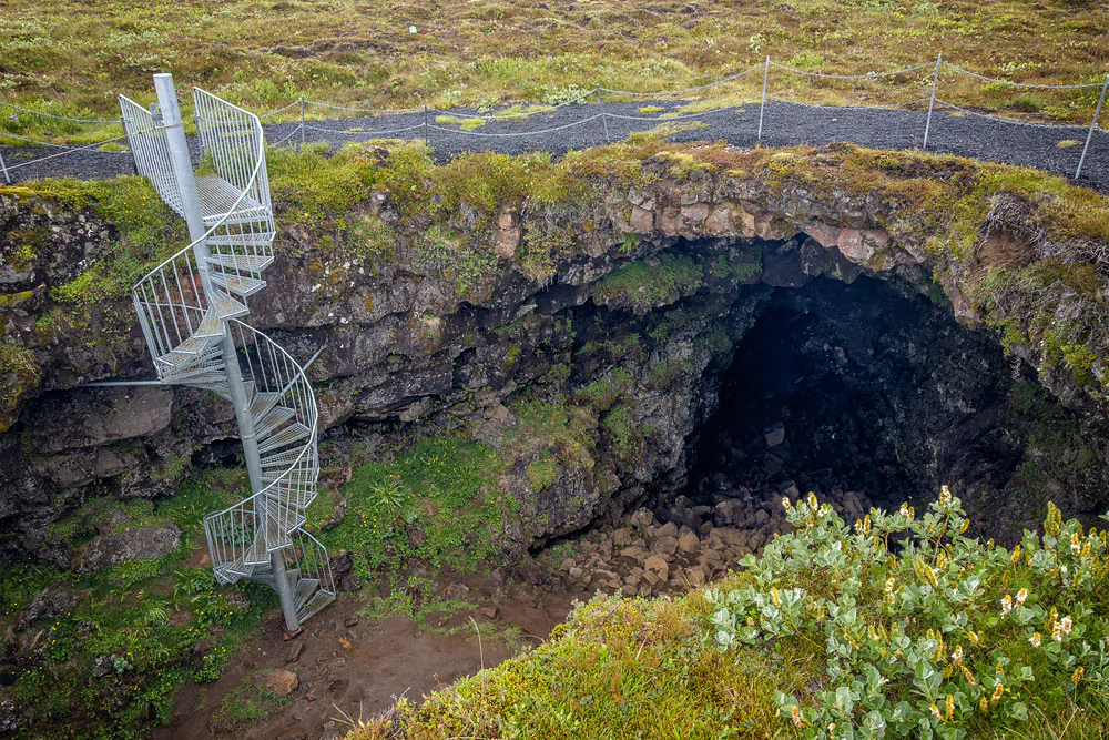Metal spiral stairs beside the collapsed Arnarker lava cave entrance in mossy Leitarhraun.